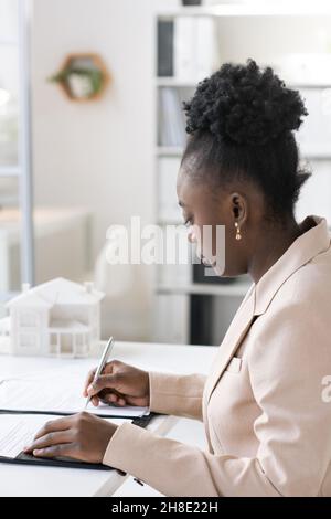 Ernsthafte junge Blackwoman mit lockigen Haaren sitzt am Tisch in der Bank und Unterzeichnung Hypothek Dokument Stockfoto