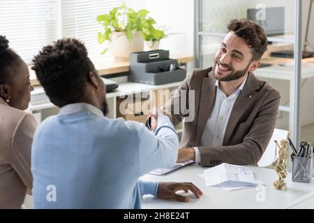 Lächelnder junger bärtiger Bankspezialist, der blackman handshake, während er das Paar im Büro begrüßt Stockfoto