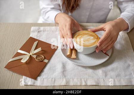 Frau mit Tasse Cappuccino auf dem Tisch im Café und Geschenkgutschein, Einladungsumschlag mit Wachsversiegelung, Hochzeitseinladungskarte. Geringe Tiefe des Fi Stockfoto