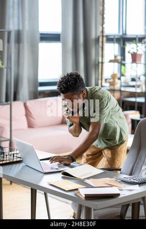 Ernsthafter junger Blackman, der am Tisch steht und Informationen auf dem Laptop überprüft, während er im Büro telefoniert Stockfoto