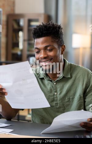 Lächelnder junger Blackman mit Bart, der im Wohnzimmer am Tisch sitzt und einen Brief von der Bank erhält Stockfoto