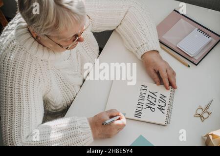 Gesunde Neujahrvorsätze für ältere Erwachsene. Ältere ältere alte Frau in weißem Pullover schriftlich handgeschriebenen Text Neujahr Auflösungen in offener Stockfoto