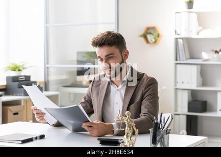 Seriöse junge bärtige Anwältin in brauner Jacke, die im Büro am Tisch sitzt und juristische Papiere liest Stockfoto