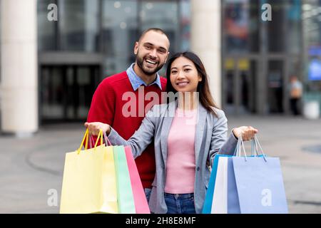 Attraktives junges, vielfältiges Paar mit bunten Geschenktüten, die in die Kamera schauen und in der Nähe eines großen Einkaufszentrums lächeln Stockfoto
