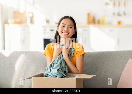 Zufriedenes Käuferkonzept. Überglücklich asiatische Frau Öffnen und Auspacken Paket nach der Lieferung, umarmende Hemd Stockfoto