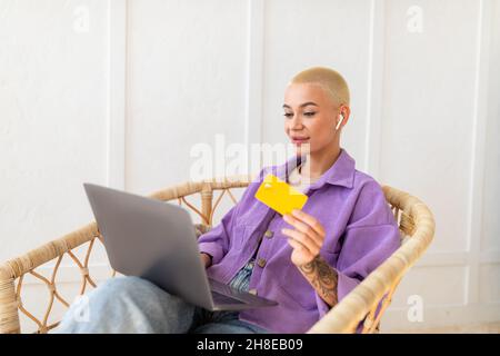 Remote-Banking. Fröhliche Frau mit Kreditkarte und Laptop, die Waren im Internet bestellt und auf einem Stuhl sitzt Stockfoto