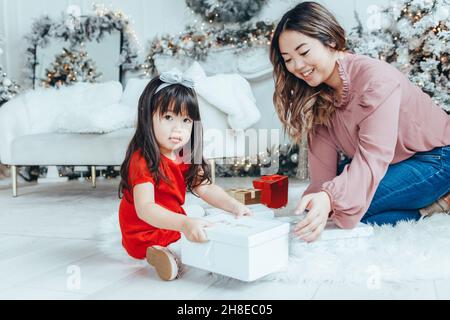 Asiatische Familie Mutter geben Geschenkbox mit Geschenk an Tochter Kleinkind Mädchen Weihnachten oder Neujahr zu feiern. Gemischte Rasse Mutter sitzt auf dem Boden mit taug Stockfoto