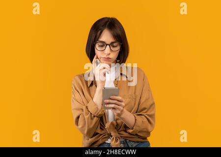 Verwirrte junge Frau, die auf das Handy schaut und denkt, die Hand in der Nähe der Wange hält und auf gelbem Studiohintergrund posiert Stockfoto