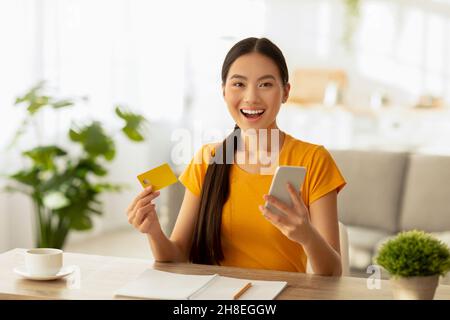 Einfaches Zahlungskonzept. Überglücklich asiatische Frau mit Kreditkarte und Handy, Bestellungen über das Internet Stockfoto