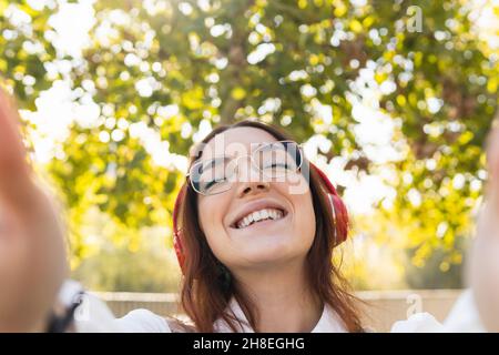 Lächelnde Rothaarige Frau mit Brille, die Selfie mit den Händen für soziale Netzwerke nimmt, während sie mit geschlossenen Augen Musik hört. Positives Konzept Stockfoto