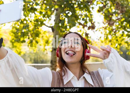 Attraktive junge Rothaarbusinessfrau mit Brille, die die Zunge zeigt und Spaß hat, während sie Selfie über das Smartphone macht und Musik im Freien hört Stockfoto
