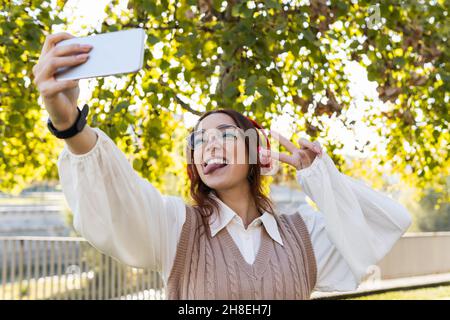 Attraktive junge Rothaarbusinessfrau mit Brille, die die Zunge zeigt und Spaß hat, während sie Selfie über das Smartphone macht und Musik im Freien hört Stockfoto