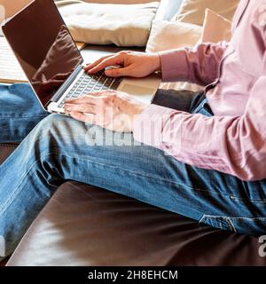Seitenansicht des anonymen Geschäftsmannes, der auf dem Sofa sitzt und auf dem Laptop tippt, während er zu Hause arbeitet Stockfoto