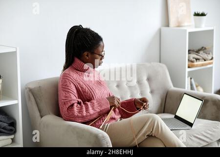 Porträt einer jungen afroamerikanischen Frau, die zu Hause strickt und einen Laptop mit leerem Bildschirm und Platz für Kopien ansieht Stockfoto