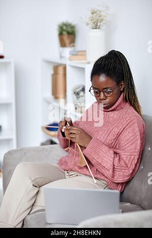 Vertikales Porträt einer jungen afroamerikanischen Frau, die zu Hause strickt und auf den Laptop-Bildschirm schaut und Bildungsvideos ansieht Stockfoto