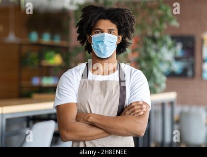 Der Junge Schwarze Barista Trägt Schürze Und Medizinische Maske Und Posiert Im Innenraum Des Cafés Stockfoto