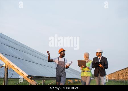 afroamerikanischer Techniker, der dem arabischen Mann und der muslimischen Frau in Hijab eine moderne Solarstation übergab. Zwei Manager mit Laptop und Clipboard steuern die Produktion von grüner Energie. Stockfoto