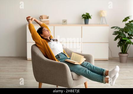 Sorgenfreie arabische Frau, die sich in einem gemütlichen Sessel ausdehnt, während sie zu Hause liest und Freizeit verbringt, um ein ruhiges Wochenende zu verbringen Stockfoto