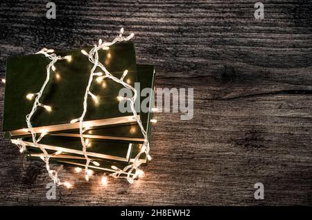 Alte Bücher, eingewickelt in Weihnachtslichter auf rustikalem dunklen Holzhintergrund. Vintage-Style. Minimaler Winterurlaub Konzept. Mit Kopierbereich. Stockfoto