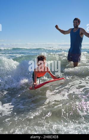 Vater beobachtete, wie der Sohn beim Boarding in der sonnigen Brandung am Meer zuschaute Stockfoto