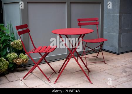 Isolierter Blick auf zwei rote Stühle an einem roten Tisch im Freien auf einer Terrasse aus Zementsteinen, mit üppigem grünem Laub im Hintergrund Stockfoto