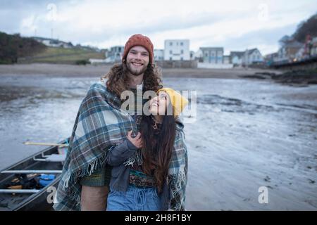 Portrait glückliches junges Paar in eine Decke gewickelt am Winterstrand Stockfoto