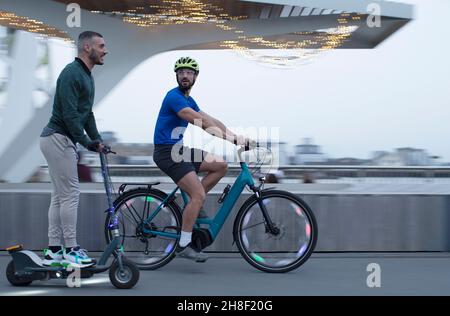 Männer reiten Roller und Fahrrad entlang der städtischen Uferpromenade Stockfoto