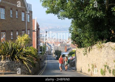 Swanage, Dorset, Großbritannien. Ein Paar, das an einem Sommermorgen einen steilen Hügel hinunter in Richtung Stadtzentrum geht, mit Blick über die Dächer zur Swanage Bay. Stockfoto