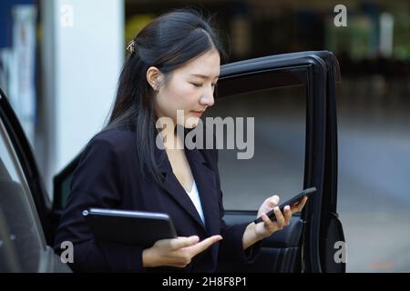 Beschäftigte asiatische Geschäftsfrau, die aus ihrem Taxi herauskommt, während sie ihre Assistentin mit einer SMS-Nachricht per Smartphone anführt. Stockfoto