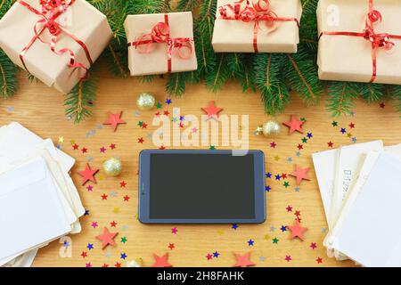 Weihnachtlicher Hintergrund mit Geschenktüten, Tablet mit schwarzem Bildschirm und Buchstaben auf einem anspruchsvollen Holztisch. Flat Lay Holiday Einkaufsliste, Happy New Year, Online-Shop, wählt Geschenke, macht Einkäufe Konzept Stockfoto