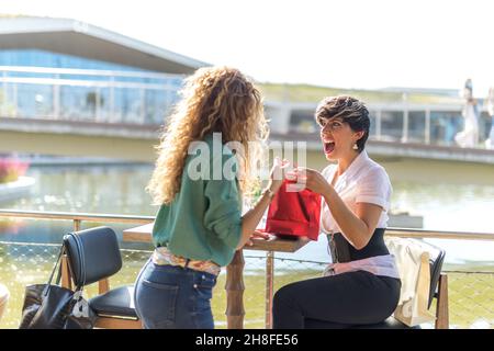 Zwei Frauen auf einer Außenterrasse. Einer von ihnen ist überrascht und freut sich über ein Geschenk des anderen. Stockfoto