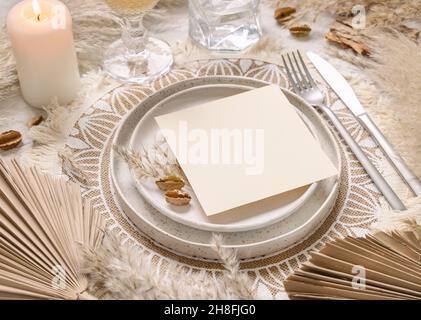 Hochzeitstisch Platz mit quadratischer unbeschriftete Karte auf böhmischen Tischset Nahaufnahme. Boho Hochzeitskarte Mockup. Tropisch beige Schablone mit getrockneten Palmblättern und Stockfoto