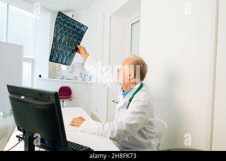 Seitenansicht eines seriös erwachsenen männlichen Arztes, der die MRT-Wirbelsäulenaufnahme eines Patienten untersucht, der am Schreibtisch mit Computer im medizinischen Bürozimmer sitzt. Stockfoto