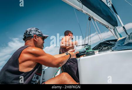 Gutaussehende starke Männer, die auf dem Segelboot arbeiten. Zieht das Seil, um den Segel zu falsen. Sommerurlaub auf dem Wasser Transport. Aktiver Urlaub. Bereisen Sie die Welt. Stockfoto