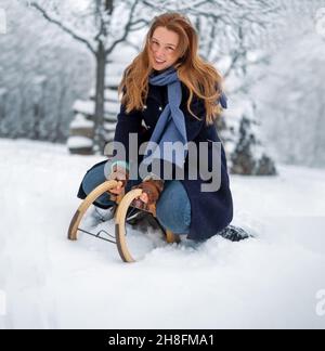 Schöne, junge, lächelnde, niedliche, rothaarige Frau mit roten Haaren, auf Schlitten sitzend, im kalten Winter frierend, ein Schal um den Hals, Kopierraum Stockfoto