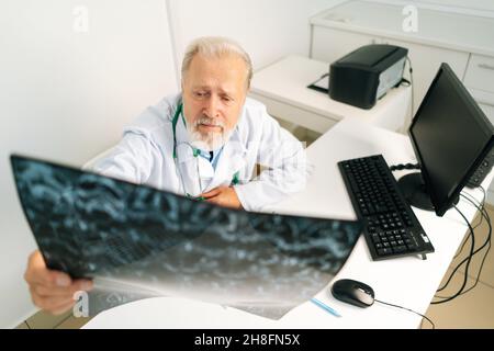 Aus der Perspektive eines erwachsenen Arztes, der die MRT-Wirbelsäulenaufnahme eines Patienten untersucht, der am Schreibtisch mit Computer im medizinischen Büroraum sitzt. Stockfoto