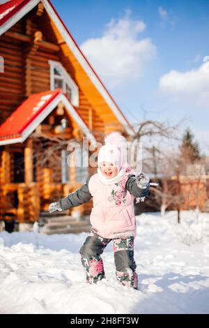 Kleines Mädchen spielt auf dem Hintergrund der Hütte. Kind hat Spaß an sonnigen Wintertag im Hinterhof in der Nähe von Holzhaus, Schneebälle spielen, springen in Stockfoto