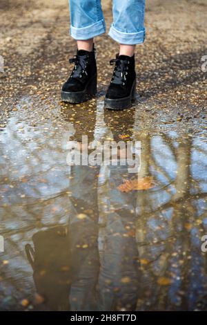 Die untere Hälfte der Beine einer Person trägt Stiefel und steht vor einer Pfütze aus Schlamm in einem Wald mit der Person und den Bäumen, die sich im Wat spiegeln Stockfoto