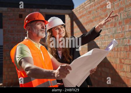 Fröhliche Architektin und ihre Partnerin schauen sich Baupläne an Stockfoto