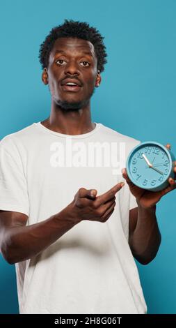 Lockere Person, die stundenlang und Minuten auf die Zeit an der Wanduhr zeigt. Porträt eines jungen Mannes, der die Uhr anschaut und den Countdown und die Verzögerung der Arbeit erklärt. Alarm für rechtzeitige Einstellung bei Erwachsenen Stockfoto