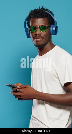 Spielerische Erwachsene tanzen im Studio zum Rhythmus singen auf Kopfhörern und blicken auf die Kamera. Fröhlicher Mann, der Musik über Smartphone und Headset hört, während er über einem isolierten Hintergrund eine Sonnenbrille trägt Stockfoto