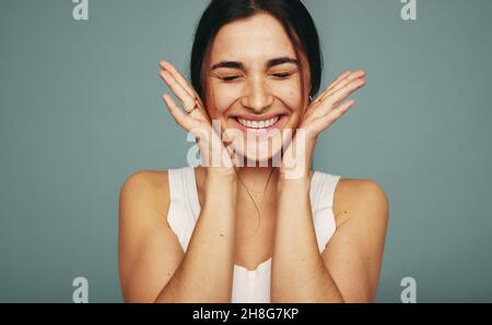 Aufgeregt junge Frau lächelt mit geschlossenen Augen in einem Studio. Glückliche junge Frau umarmt ihr Gesicht, während sie allein vor einem Studiohintergrund steht. Stockfoto