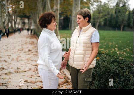 Zwei ältere Frauen lächelten sich gegenseitig. Zwei alte Freunde trafen sich im Park. Aktiver Lebensstil von älteren und Frauen mittleren Alters. Herbstliches kühles Wetter Stockfoto