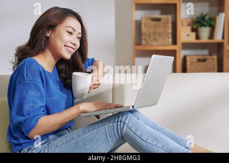 Lächelnde junge asiatische Frau, die mit einem Laptop auf den Knien sitzt und Kaffee trinkt, während sie zu Hause arbeitet Stockfoto