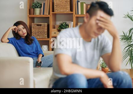 Wütende junge asiatische Frau, die mit dem Telefon auf dem Sofa sitzt und auf den Freund schaut, der das Gesicht mit der Hand bedeckt, Streitkonzept Stockfoto