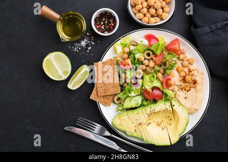 Gesunder veganer, ausgewogener Essenssalat mit Kichererbsen-Hummus, Avocado, Tomaten und grünem Salatblatt. Kopiebereich für schwarzen Hintergrund von oben Stockfoto