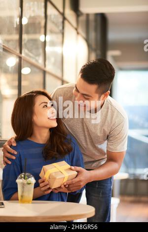 Lächelnder junger Mann umarmt asiatische Freundin von hinten im Café und gibt ihr ein Geschenk Stockfoto