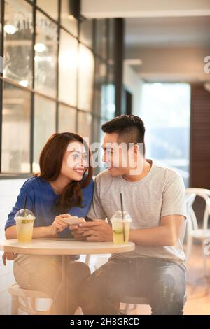 Lächelndes, schönes junges asiatisches Paar, das an einem kleinen runden Tisch mit erfrischenden Cocktails im Café sitzt und durch das Smartphone scrollt Stockfoto