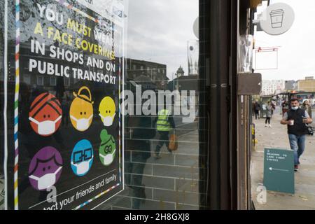 London, Großbritannien, 30. November 2021: In Clapham sind Käufer nun gesetzlich verpflichtet, in Geschäften oder öffentlichen Verkehrsmitteln eine Gesichtsmaske zu tragen. In einem Schaufenster erinnert ein buntes Schild mit einer Vielzahl von Menschen in Gesichtsmasken, geliefert vom Lambeth-Rat, an die heute in Kraft getretene Änderung der Vorschriften. Anna Watson/Alamy Live News Stockfoto