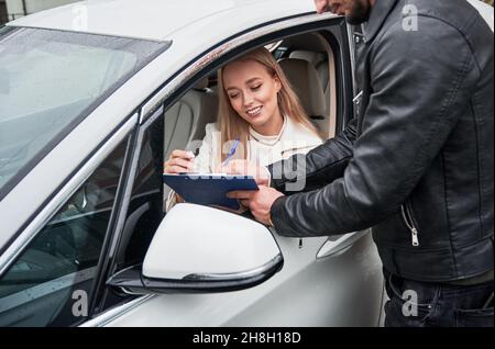 Die blonde Geschäftsfahrerin sitzt auf dem Fahrersitz im Auto und lernt die Dokumente aus dem Klemmbrett kennen, die ihr der Mann durch das abgesenkte Fenster in der Seitentür vorgelegt hat. Stockfoto